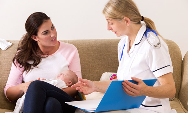 nurse sits on sofa talking to woman who is holding an infant