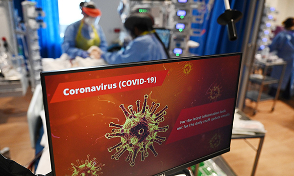 Picture of COVID-19 notice on a glass door. In the background clinical staff wearing personal protective equipment look after a patient in an intensive care unit.