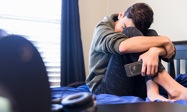 Picture of a boy sitting on a bed and pressing his face against his knees.
