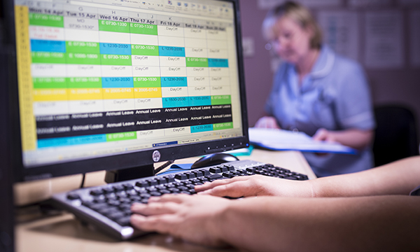 Picture shows a computer screen displaying a work rota, with a nurse in the background