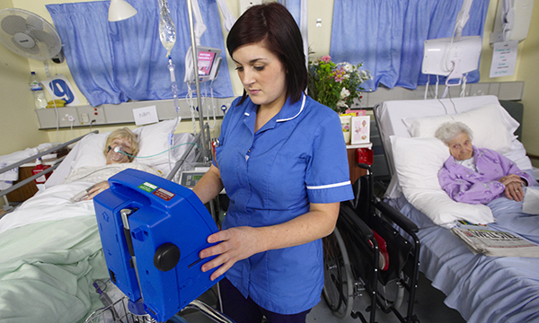 A nurse checking a monitor on a hospital ward, with two patients in the background