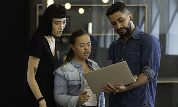 Two women and a man share and discuss ideas standing in a huddle around a laptop