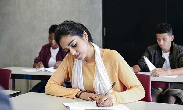 A young woman sits at a desk in a test centre, with two others at desks behind her, taking a language test for internationally trained nurses