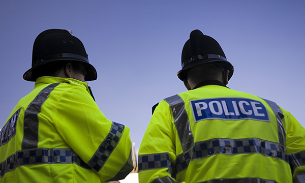 Two male police officers wearing Custodian police helmets and high vis reflective jackets in the street, their backs to the camera and looking at an incident
