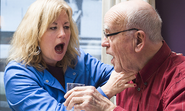 A speech and language therapist assists a patient to drink a glass of water, gently touching his throat while encouraging him to swallow by mimicking the action