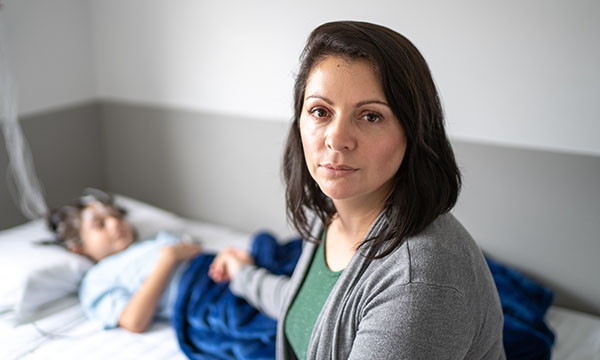 A mother, looking towards the camera, holds the hand of her child who is lying in bed