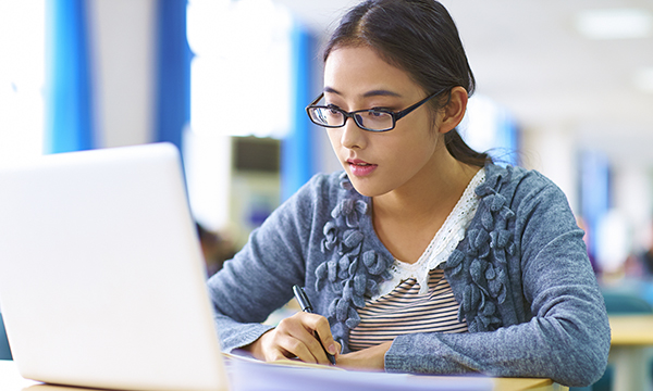 A student looks at an open laptop with pen in her hand to take notes
