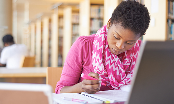 A nursing student makes notes on paper while looking at the page and her laptop computer