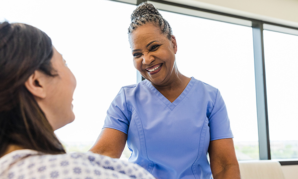 Nurse smiles broadly as she greets female patient who is lying in hospital bed