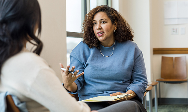 A woman sits and explains something to another woman as she holds a pen and a clipboard
