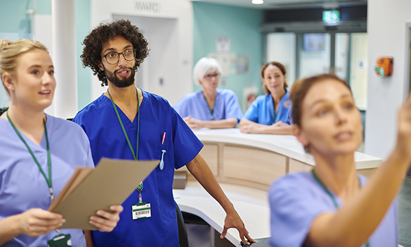 Two nurses in an emergency department look on as another nurse writes on the care planning board at the nurses’ station