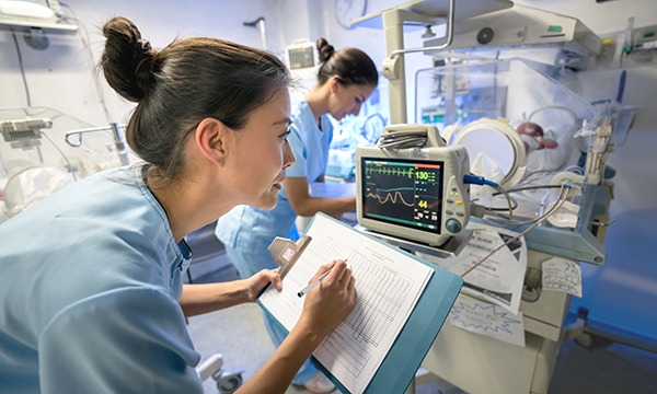 A nurse makes notes on a chart while looking at a medical monitor attached to an infant’s bed