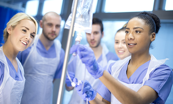 Five nursing students stand in semi-circle. They watch as one handles IV line – NMC is to review undergraduates clinical learning requirements