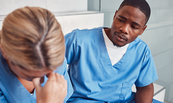 A nurse holds a hand to her forehead in apparent distress as a colleague looks on anxiously