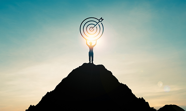 Illustration showing a figure standing at the top of a peak highlighted against a golden sky and holding up a target with an arrow through the centre, suggesting reaching a goal