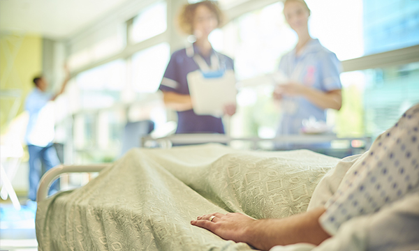 Two nurses stand looking at a patient lying in a hospital bed, discussing discharge plans