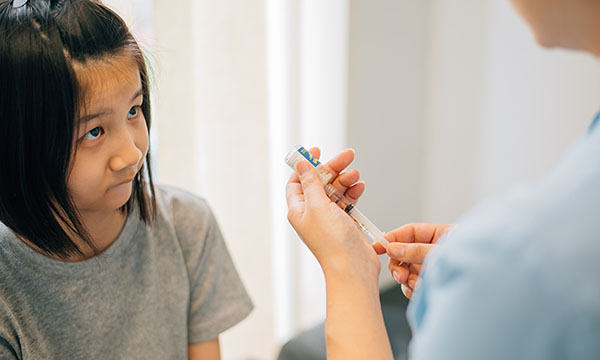 A young girl looks apprehensive as she watches a nurse preparing an injection