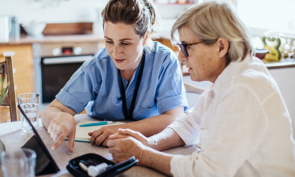 A healthcare professional and older patient run through a blood monitoring process with the aid of a tablet computer at the patient’s home