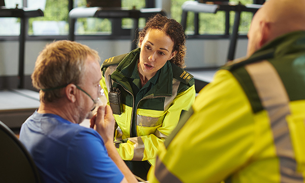 An ambulance nurse wearing a high visibility jacket talks to a patient who is holding an oxygen mask to his face