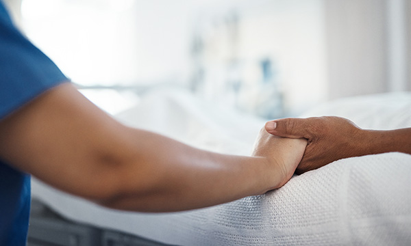 Close-up image of two hands, as a nurse in uniform sits at the bedside of a dying patient in hospital, holding their hand