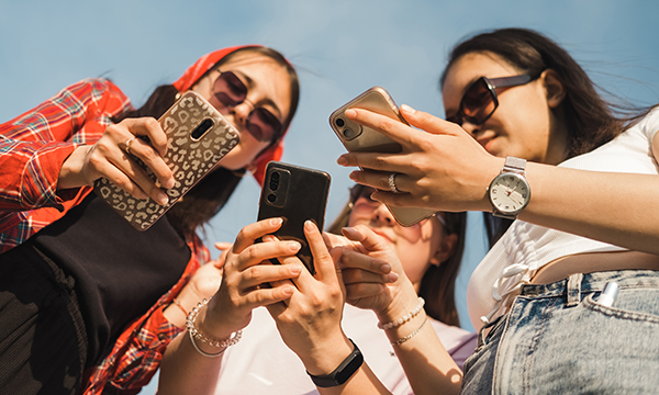 Three young woman huddle together, all looking at their phones. Social media can effective in advertising nurse vacancies