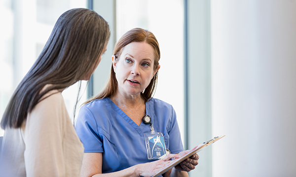 A patient, seen from behind, talks in confidence to a nurse in uniform who is holding a clipboard