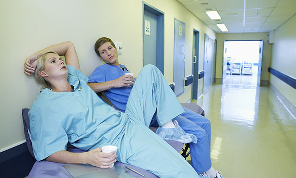 Two weary emergency care nurses, both holding disposable paper cups, rest on a trolley in a hospital corridor