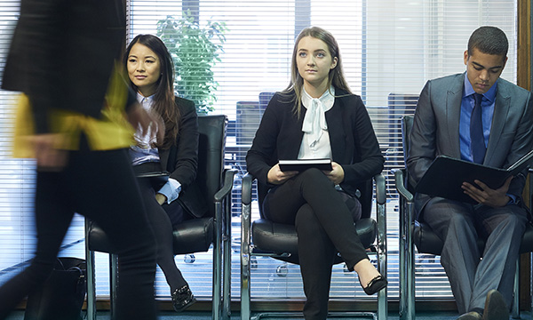 A row of people in smart attire seated in a waiting area looking slightly anxious and preoccupied, as if waiting to be called into a job interview