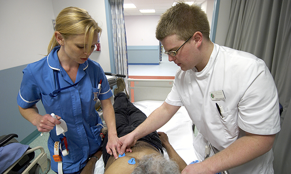 Nursing student is mentored by a nurse as he places ECG leads on the chest of a prone patient