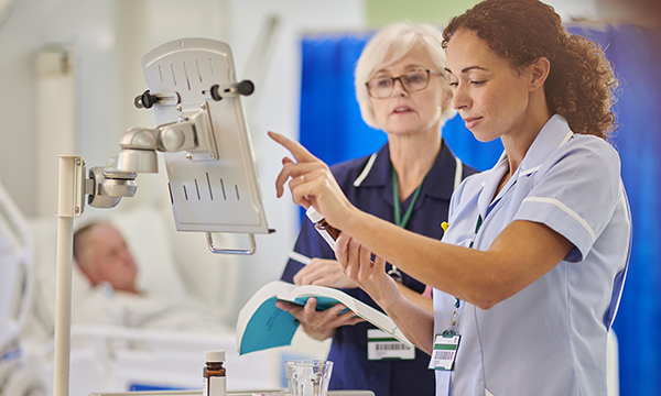 Two nurses, one holding a reference book and the other a bottle of medicine, consult a display board in a ward setting