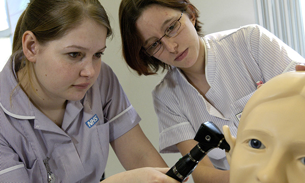 A nursing student holds an otoscope to the ear of a manikin as her trainer looks on