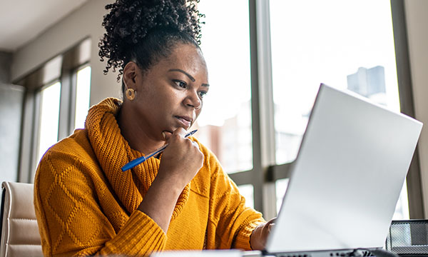 A woman sits at a desk with a laptop in front of her, looking thoughtful as she reads what is on the screen, as if preparing a supporting statement for a job application