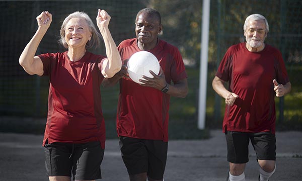 An older woman celebrates a walking football victory with her two male teammates (one of whom is holding a football): activities like this can be an important part of healthy ageing