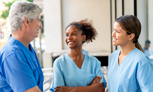 Three nurses stand talking happily. Two are younger, one is older, illustrating intergenerational teamwork in nursing
