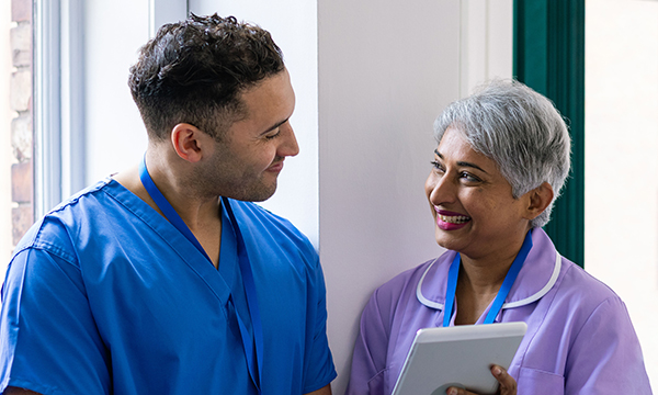 A smiling senior nurse chats to a younger colleague and shares information via a tablet computer