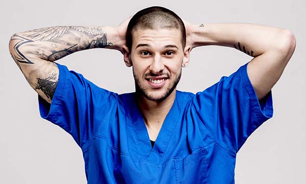 A nurse in scrubs with tattoos on both arms and a pierced ear, standing in a relaxed pose with his hands behind his head