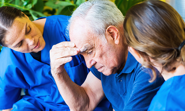 An older man with his head bowed appears pensive as he holds a hand to his forehead, while nurses sitting either side of him watch closely and try to engage him in conversation 