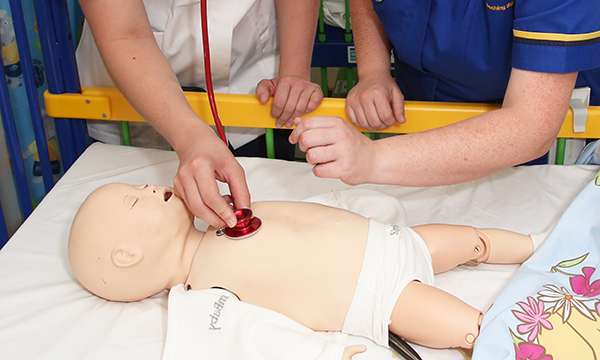 A manikin of an infant lying on a bed is used to help a nursing student practise using a stethoscope