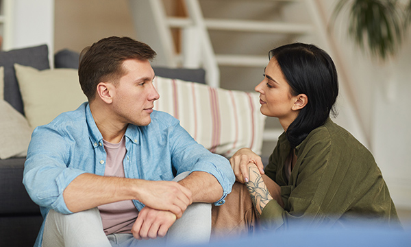 Heterosexual couple in their thirties sitting on the floor and looking at each other with relaxed expressions but crossed arms, suggesting an attempt at communication 