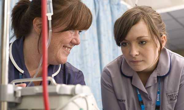 A nursing student looks at an IV monitor as she listens to a senior nurse while learning on placement