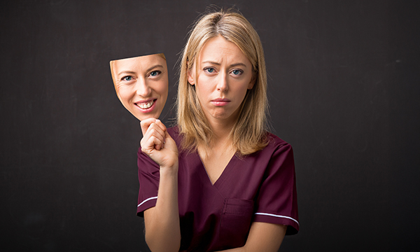 People pleasing can take a toll on your well-being: a nurse holds a smiling mask of her face away from her real face, which looks sad  