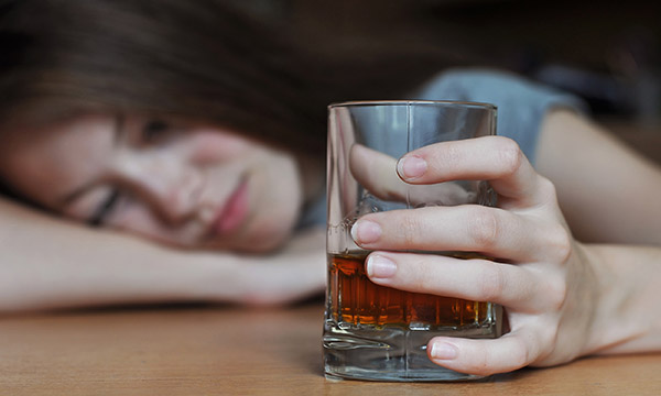 Alcohol addiction among nurses: a sad looking young woman at a desk with her head on her arm, holding a glass containing alcohol