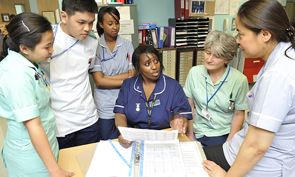 A ward manager sits in an office in a clinical setting holding a staff rota as she talks to her nursing team who are standing around her
