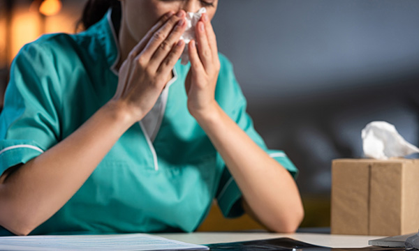 Nurse sickness: a nurse sitting at a desk at work holds a tissue to her face, as if blowing her nose
