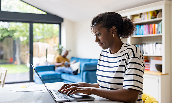 Written reflective account: a woman sits at a table in her living room typing on a laptop