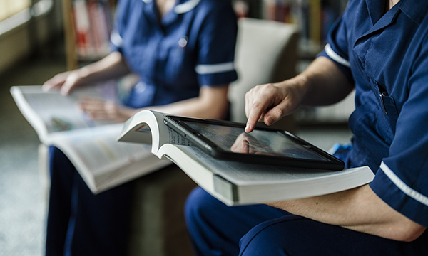 Reflective practice: two nurses seated in a library, each with a book, one has an iPad too