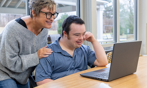 Learning disability nursing must be positioned as a compassionate profession: a nurse guides a service user who is happily sitting in front of a laptop at a desk&nbsp;
