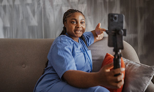 A nurse sitting on a sofa looks into a camera while creating a video. Social media posts are about making a difference for the people who need nurses most.