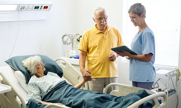 Obtaining a clear background history is essential, particularly in the presence of confusion, falls or social-related concerns. Picture shows a nurse speaking to an older woman lying in a hospital bed and an older relative holding their hand