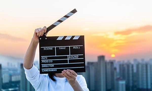 A person holds a clapper board on a film set, standing at the top of a building overlooking a city skyline